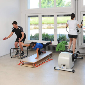 Trois personnes s'entraînent dans une salle lumineuse. À gauche, un homme effectue des exercices avec des barres parallèles. Au centre, une femme est allongée sur une plate-forme, exécutant un mouvement de renforcement musculaire. À droite, une autre femm