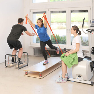 Image montrant trois personnes s'entraînant dans une salle de sport. Une femme en t-shirt bleu utilise des bandes de résistance, tout en s'équilibrant sur une plateforme. À côté, un homme en noir fait des exercices de renforcement, et une autre femme en t