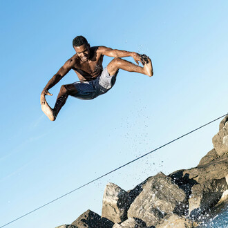 Jumping and holding his feet in the air while above a slackline. Water is spashing up from the ocean.