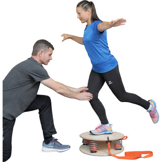 Un homme aide une femme à s'équilibrer sur un équipement de fitness en bois avec des ressorts. La femme, en tenue de sport bleu et leggings noirs, est en position de déséquilibre, les bras tendus. L'homme, en vêtement gris, est accroupi et soutient la jam