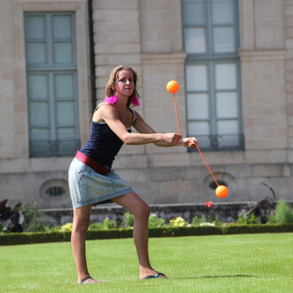 Femme jonglant avec des bolas orange, portant un débardeur noir et une jupe en jean, dans un jardin bien entretenu avec des fleurs et des bâtiments en arrière-plan.