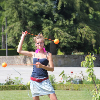 Jeune femme en train de jongler avec des bolas contact de 80 mm, portant un débardeur noir et une jupe claire. Elle a des plumes fuchsia dans les cheveux et utilise des cordes orange pour faire tournoyer les bolas dans un jardin verdoyant.