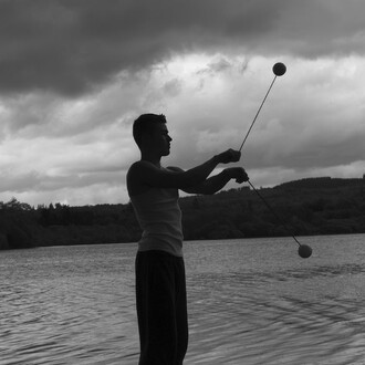 Silhouette d'un jongleur utilisant des bolas contact de 80 mm au bord d'un lac, avec un ciel nuageux en arrière-plan.