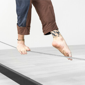 Image montrant des pieds nus marchant sur un cable de fil d'équilibre. On voit une partie de la structure métallique noire en arrière-plan. L'image met en avant l'utilisation de la slackline pour l'exercice d'équilibre.