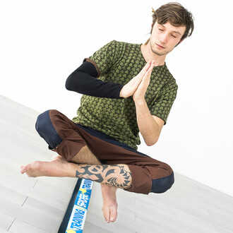 Image montrant une personne assise en position de méditation sur une slackline bleue, les mains jointes devant la poitrine. Elle porte un t-shirt vert à motifs et un pantalon marron. L'image illustre la stabilité et le contrôle nécessaires pour maintenir 