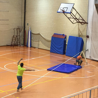 
personne pratiquant la slackline dans un gymnase, avec des tapis de protection et des équipements sportifs en arrière-plan. L'ancrage se fait ici avec le plateau dans le mur et la sangle passe sur un triangle de bois.