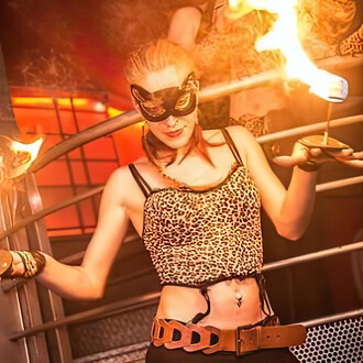 Artiste de feu exécutant une performance spectaculaire avec des mains de feu, vêtue d'un costume léopard et portant un masque. La danseuse tient des torches enflammées sur ses paumes, illuminant la scène avec des effets visuels captivants. L'ambiance est 