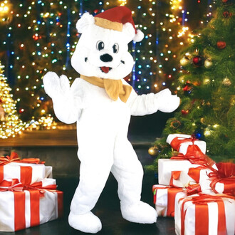 Mascotte ours de Noël en costume blanc avec un chapeau de Santa, se tenant dans un décor festif entouré de sapins illuminés et de cadeaux décorés.