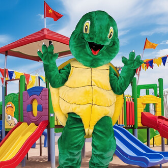 Mascotte en costume de tortue souriante, avec un corps vert et une carapace jaune, se tenant devant une aire de jeux colorée avec des toboggans rouges et bleus, des drapeaux multicolores et un ciel bleu.