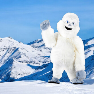 Mascotte Yeti se tenant debout sur un terrain enneigé, avec des montagnes enneigées en arrière-plan. Il est entièrement recouvert de fourrure blanche, avec un visage souriant et des yeux bleus. Il lève une main, en faisant un signe amical.