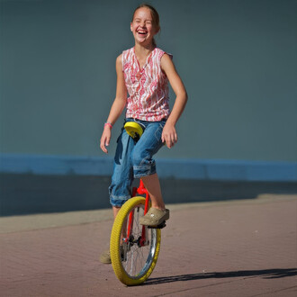 Fille souriante en train de faire du monocycle Quax Luxus 20, portant un haut à motifs et un short en denim, sur un chemin ensoleillé.