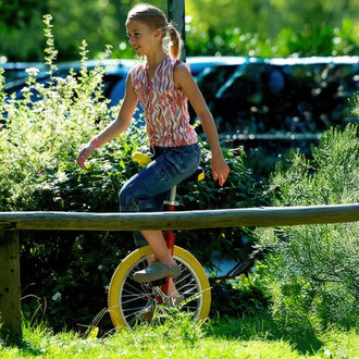 Une jeune fille souriante fait du monocycle Quax Luxus 20 sur une barrière en bois. Elle porte un haut coloré et un pantalon en jean. En arrière-plan, on distingue des plantes vertes et un environnement naturel ensoleillé.