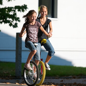 Deux filles joyeuses se tiennent sur un monocycle, souriant et s'amusant. L'une porte un t-shirt gris avec un motif de hibou, tandis que l'autre est vêtue d'un haut à pois noir. Le monocycle a une grande roue jaune à l'avant et une petite roue blanche à l