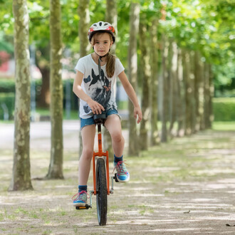 Fille sur un monocycle Quax Luxus 20 orange, portant un casque et un t-shirt avec un motif de chien, dans un cadre de parc avec des arbres alignés.