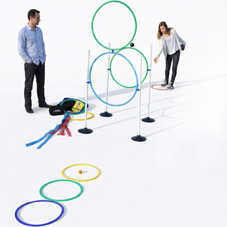 Kit d’initiation pétanque: ensemble d’entraînement avec cercles etanneaux colorés (bleu, vert et jaune) montés sur poteaux, petit ballon orange au sol, sacs et rubans colorés à gauche, deux personnes observant près du dispositif.