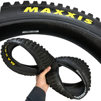 Pneu 20x2.5 pour vélo de cross, avec une main tenant deux pneus distincts, le tout sur un fond blanc. L'un des pneus est enroulé en arrière-plan et porte le logo MAXXIS en jaune, tandis que les deux pneus tenus main sont également marqués MAXXIS et Quax. 