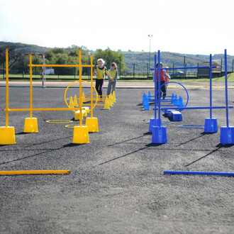 Jeux d'agilité en extérieur avec des obstacles colorés en jaune et bleu, incluant des cerceaux et des barrières, utilisés par des enfants déguisés en gilets. En arrière-plan, uneInstallation sportive et des collines verdoyantes sous un ciel ensoleillé.