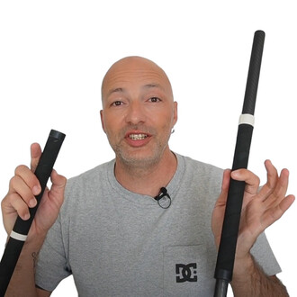 Un homme souriant tenant deux tubes en carbone pliables, l'un dans chaque main. Le tube de droite est plus long que celui de gauche. L'arrière-plan est blanc, mettant en valeur les tubes. L'homme porte un t-shirt gris avec le logo DC.