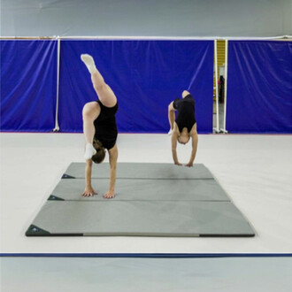 Deux gymnastes effectuent des exercices sur le Tapis Housse Evolution 40, avec une gymnaste exécutant un handstand et l'autre en transition. Le tapis est disposé sur un sol clair, et une paroi bleue est visible en arrière-plan.